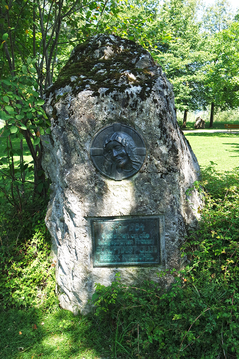 Gedenkstein aus Dolomitgestein mit rechteckiger Bronzetafel mit Inschrift und kreisrundem Madonnenrelief an der Redfordpromenade in St. Johann in Tirol.